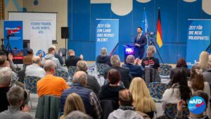Ein Bürgerdialog der AfD-Westerwald in der Götzenberg-Halle. Dennis Hohloch steht am Podium vor sitzendem Publikum. Hinter ihm hängen blaue Plakate mit politischen Slogans der AfD und die deutsche Flagge. Auf der linken Seite sind Banner der Partei AfD zu sehen. Die Zuhörerinnen und Zuhörer sitzen in Reihen und applaudieren.