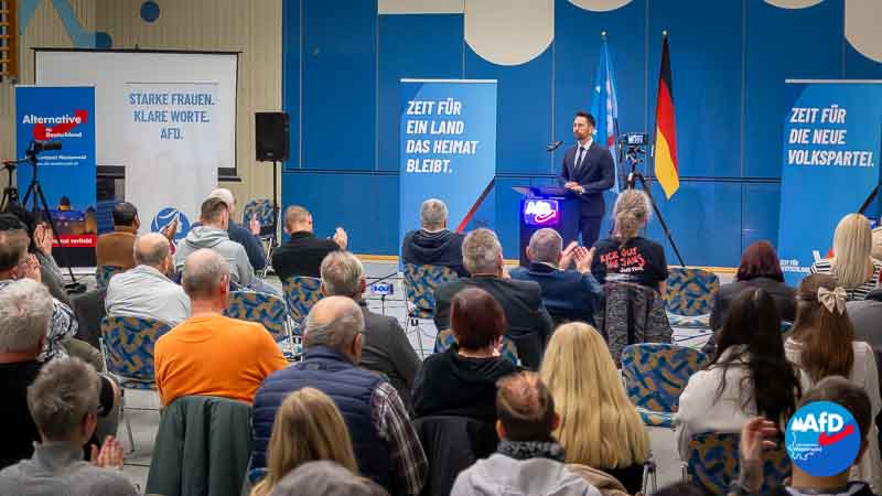 Ein Bürgerdialog der AfD-Westerwald in der Götzenberg-Halle. Dennis Hohloch steht am Podium vor sitzendem Publikum. Hinter ihm hängen blaue Plakate mit politischen Slogans der AfD und die deutsche Flagge. Auf der linken Seite sind Banner der Partei AfD zu sehen. Die Zuhörerinnen und Zuhörer sitzen in Reihen und applaudieren.