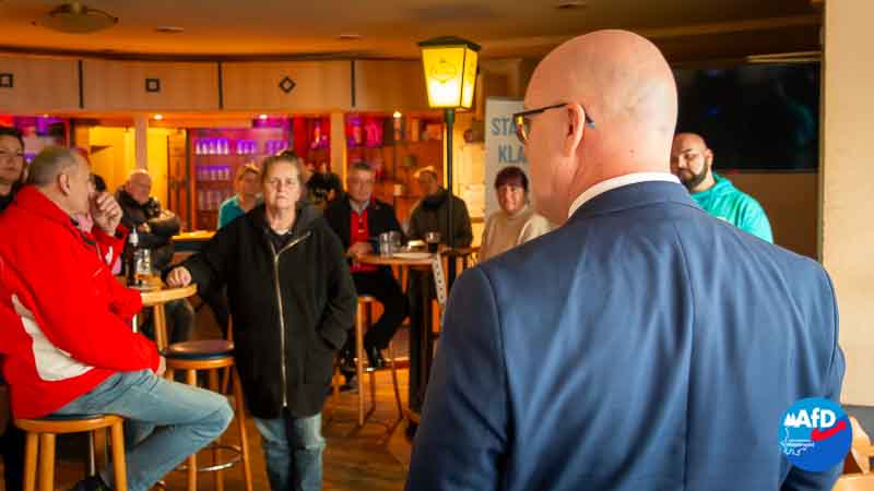 In einer Gaststätte spricht ein Mann im Anzug mit dem Rücken zur Kamera zu einer kleinen Gruppe von Menschen, die an Tischen und Barhockern sitzen und aufmerksam zuhören. Im Hintergrund ist die Einrichtung einer Bar zu sehen sowie ein Banner mit politischem Slogan.
