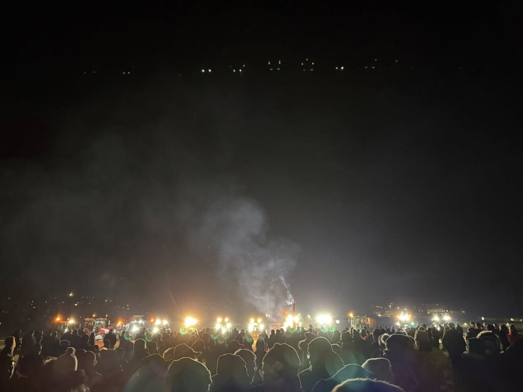 Nächtliches Mahnfeuer gegen Wolfsrisse in Rehe im Westerwald: Viele Menschen stehen dicht gedrängt im Dunkeln, im Hintergrund erhellen Feuer und Fahrzeuge den Platz, Rauch steigt in den Nachthimmel.