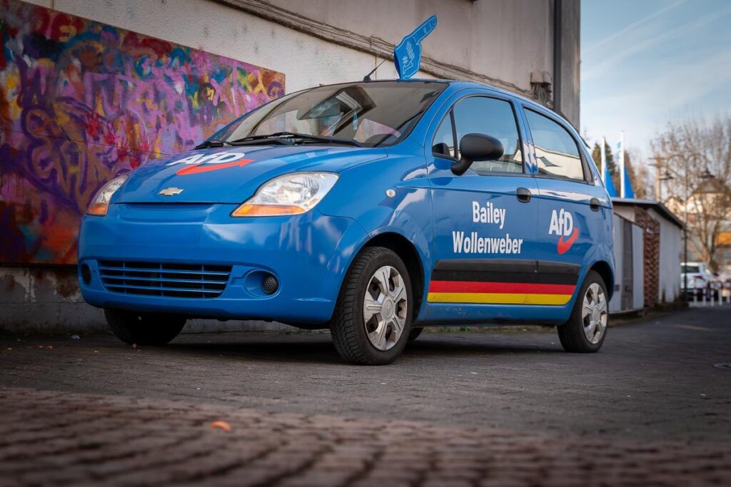 Blaues Kampagnenfahrzeug von Bailey Wollenweber, Landtagskandidat für den Wahlkreis 5, mit AfD-Logo und Deutschlandstreifen, geparkt in urbaner Umgebung in der Nähe des Marktplatzes in Selters/Westerwald