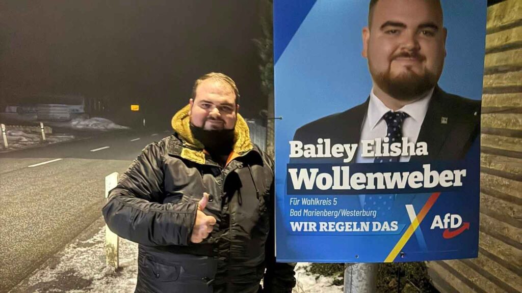 Bailey Wollenweber, Landtagskandidat der AfD Westerwald, steht abends am Straßenrand im winterlichen Westerwald neben seinem Wahlplakat und zeigt den Daumen nach oben.