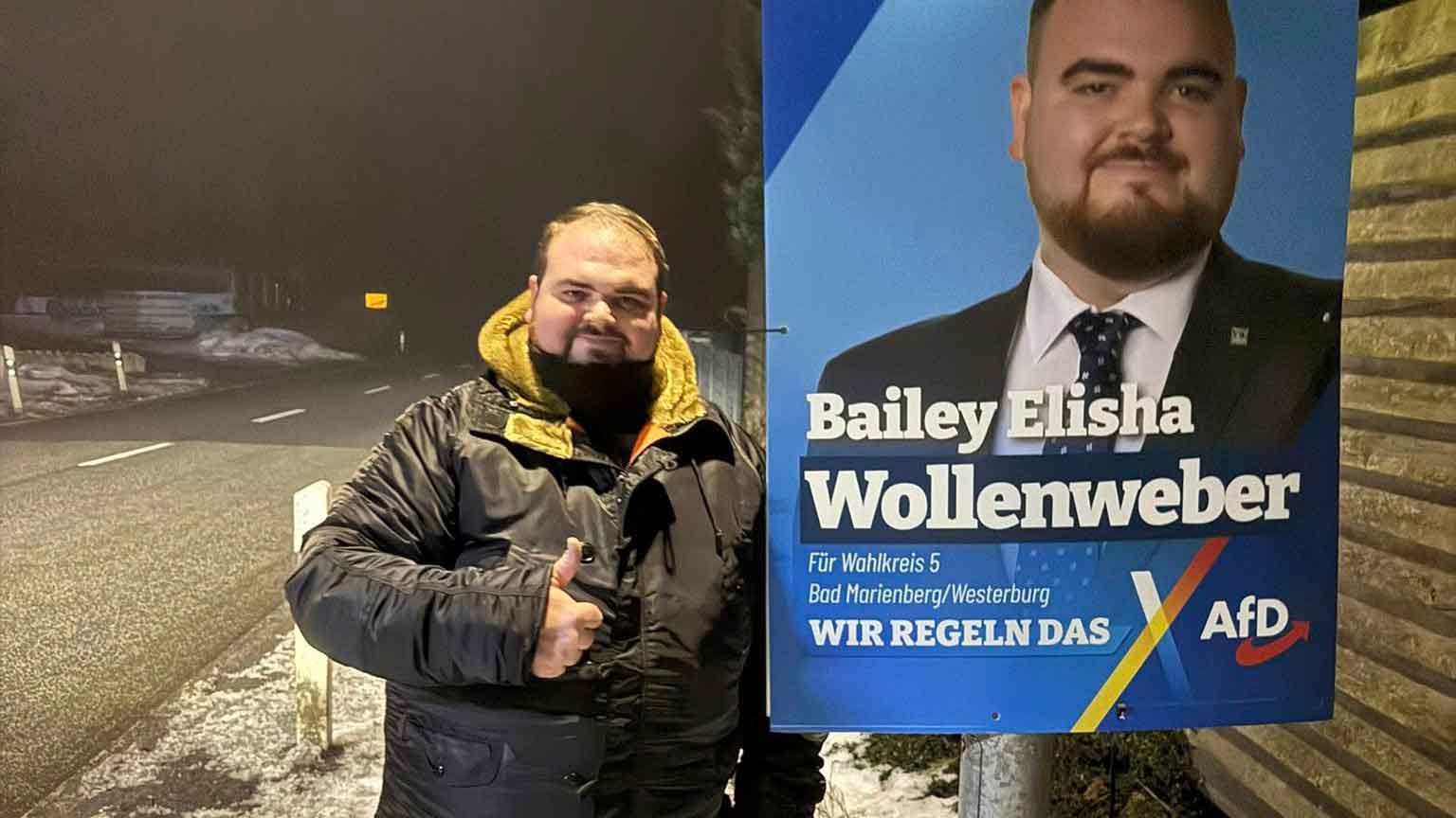 Bailey Wollenweber, Landtagskandidat der AfD Westerwald, steht abends am Straßenrand im winterlichen Westerwald neben seinem Wahlplakat und zeigt den Daumen nach oben.
