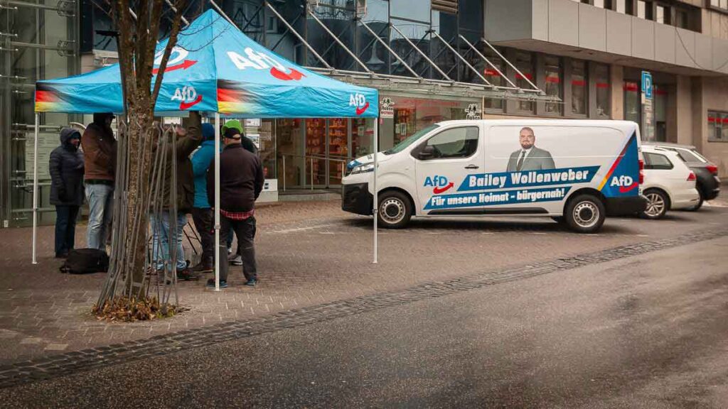 Infostand der AfD im Westerwald: Mehrere Personen stehen unter einem blauen AfD-Pavillon, daneben ein AfD-Wahlkampfbus mit dem Porträt von Bailey Wollenweber in einer Fußgängerzone.