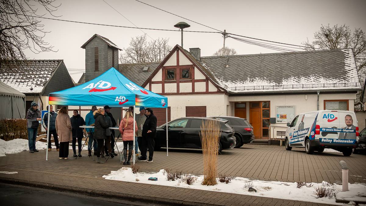 Infostand der AfD Westerwald mit blauem Pavillon, mehreren Bürgern im Gespräch und einem Wahlkampf-Fahrzeug vor einem Dorfgemeinschaftshaus im winterlichen Ortskern.